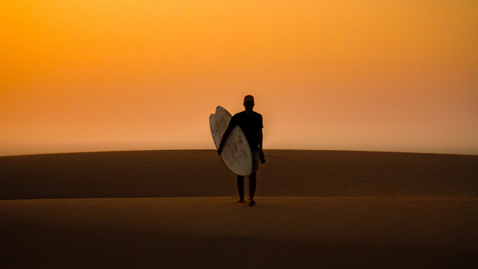 Surfer walking across the sand at sunset carrying a surfboard